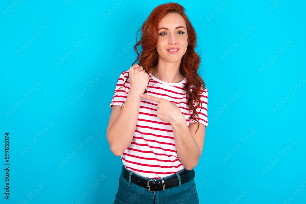Fototapeta premium young redhead woman wearing striped T-shirt over blue background In hurry pointing to wrist watch, impatience, looking at the camera with relaxed expression
