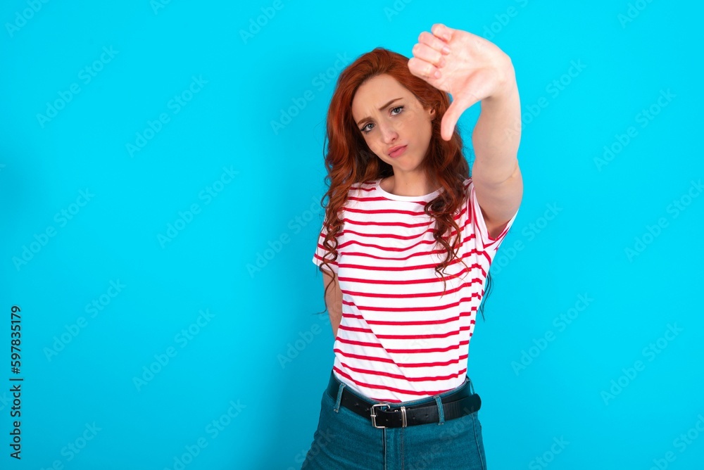 young redhead woman wearing striped T-shirt over blue background feeling angry, annoyed, disappointed or displeased, showing thumbs down with a serious look young redhead woman wearing striped T-shirt over blue background feeling angry, annoyed, disappointed or displeased, showing thumbs down with a serious look