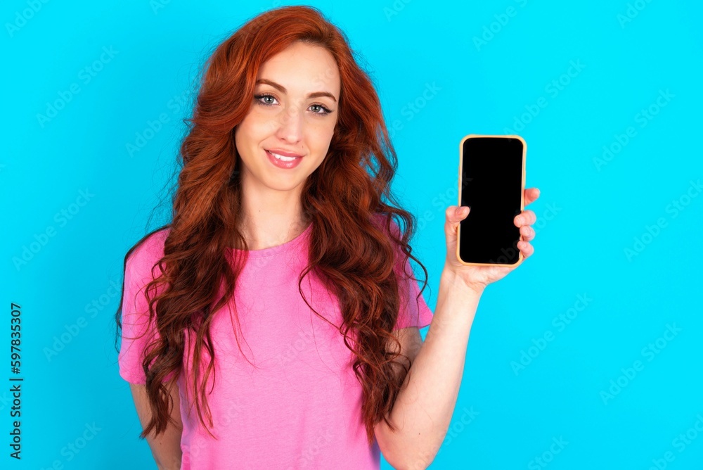 Smiling young redhead woman wearing pink T-shirt over blue background Mock up copy space. Hold mobile phone with blank empty screen Smiling young redhead woman wearing pink T-shirt over blue background Mock up copy space. Hold mobile phone with blank empty screen
