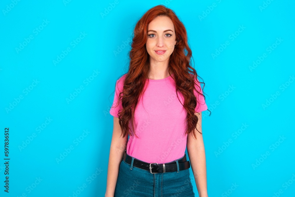 young redhead woman wearing pink T-shirt over blue background with nice beaming smile pleased expression. Positive emotions concept young redhead woman wearing pink T-shirt over blue background with nice beaming smile pleased expression. Positive emotions concept