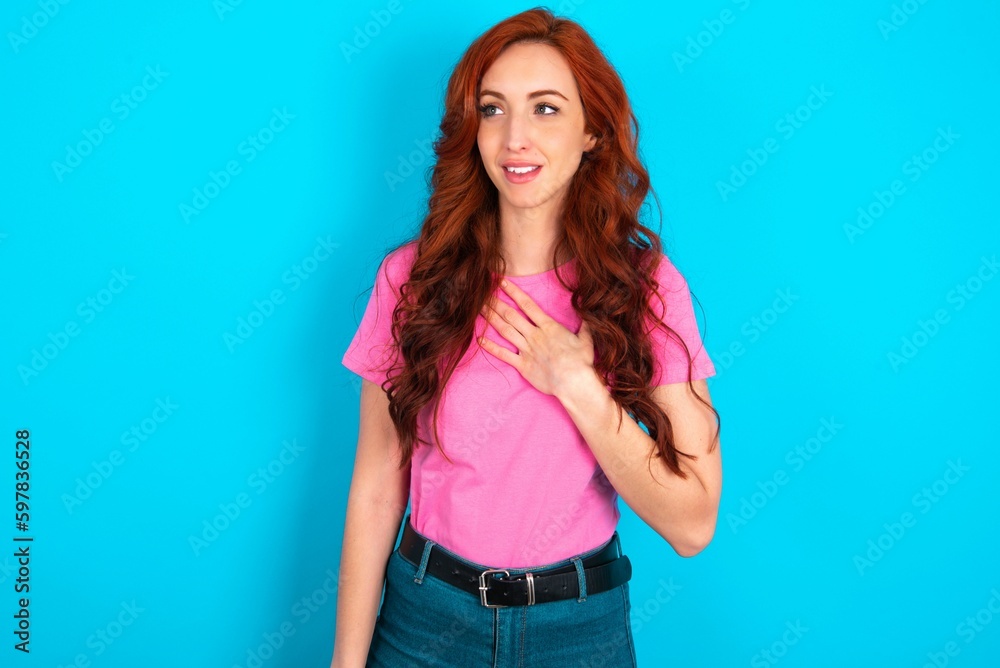 Joyful young redhead woman wearing pink T-shirt over blue background expresses positive emotions recalls something funny keeps hand on chest and giggles happily. Joyful young redhead woman wearing pink T-shirt over blue background expresses positive emotions recalls something funny keeps hand on chest and giggles happily.