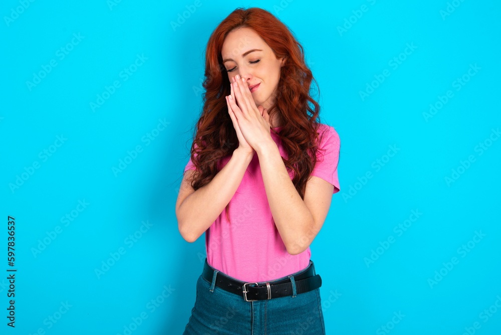 Overjoyed young redhead woman wearing pink T-shirt over blue background laughs joyfully and keeps palms pressed together hears something funny Overjoyed young redhead woman wearing pink T-shirt over blue background laughs joyfully and keeps palms pressed together hears something funny