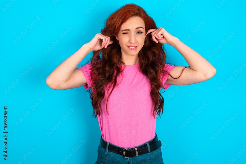 Happy young redhead woman wearing pink T-shirt over blue background ignores loud music and plugs ears with fingers asks to turn off sound