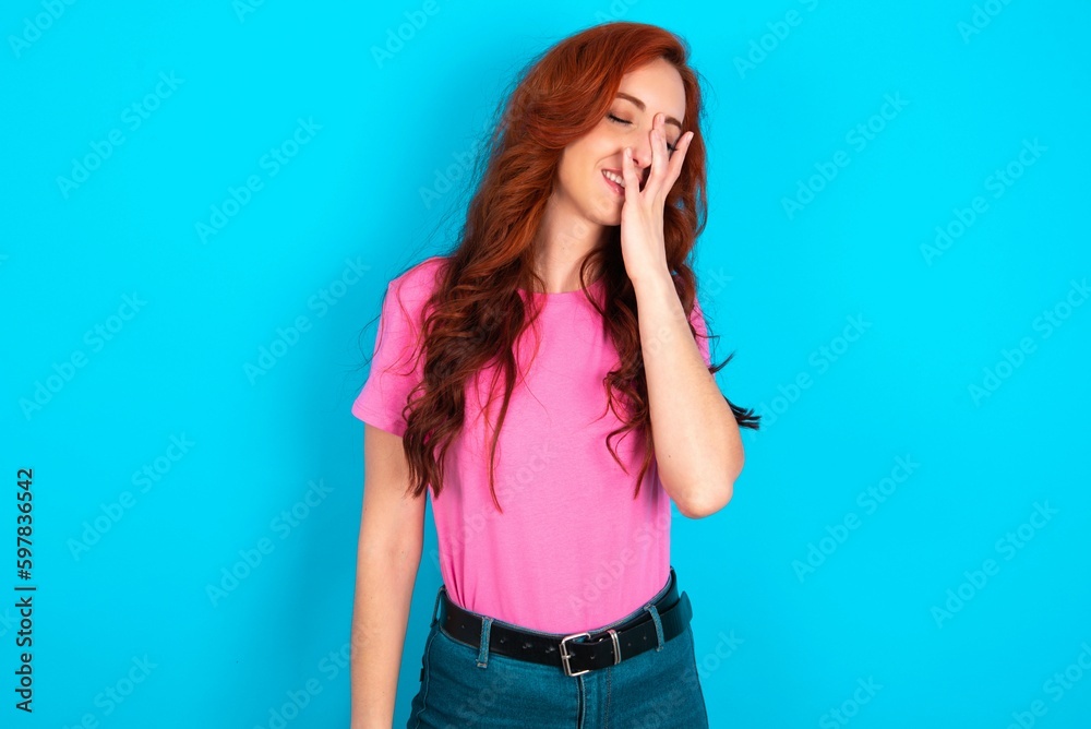 young redhead woman wearing pink T-shirt over blue background makes face palm and smiles broadly, giggles positively hears funny joke poses young redhead woman wearing pink T-shirt over blue background makes face palm and smiles broadly, giggles positively hears funny joke poses