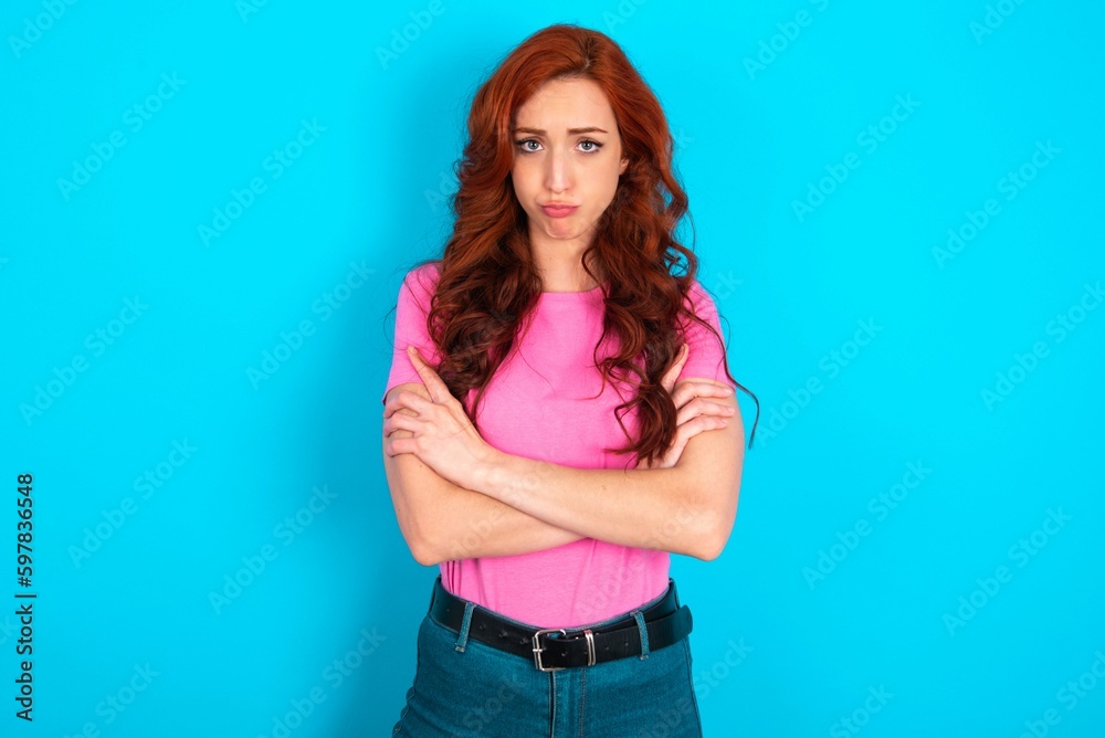 Gloomy dissatisfied young redhead woman wearing pink T-shirt over blue background looks with miserable expression at camera from under forehead, makes unhappy grimace Gloomy dissatisfied young redhead woman wearing pink T-shirt over blue background looks with miserable expression at camera from under forehead, makes unhappy grimace