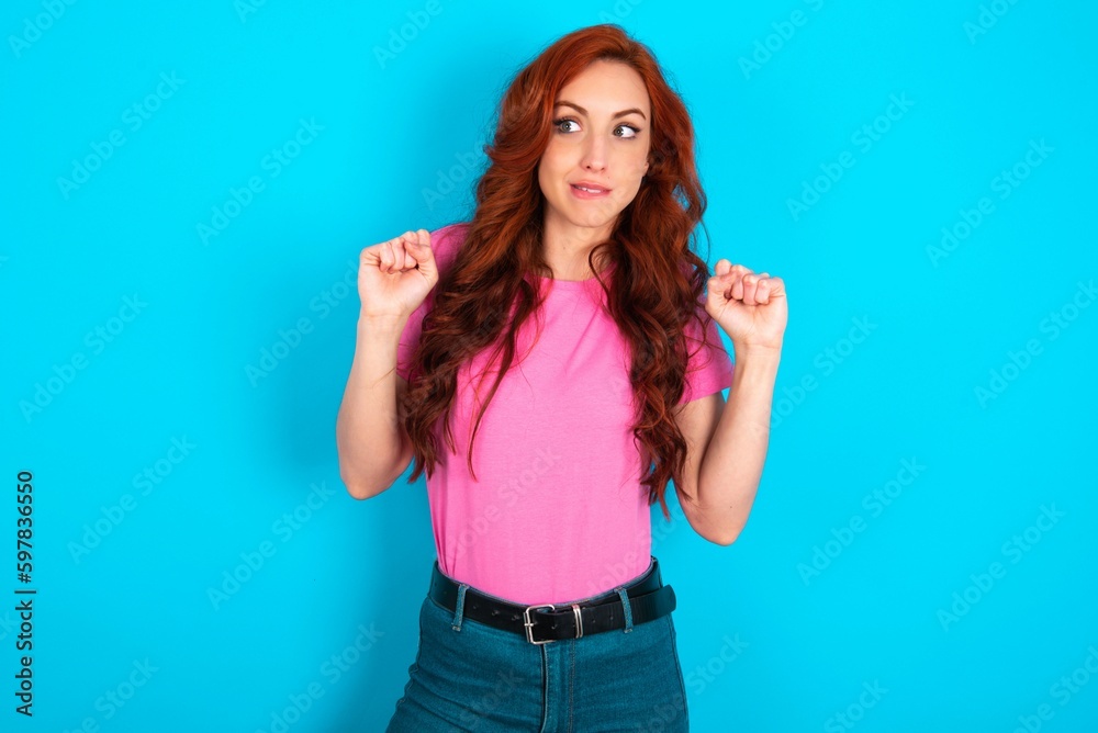Fototapeta premium young redhead woman wearing pink T-shirt over blue background clenches fists and awaits for something nice happened looks away bites lips and waits announcement of results