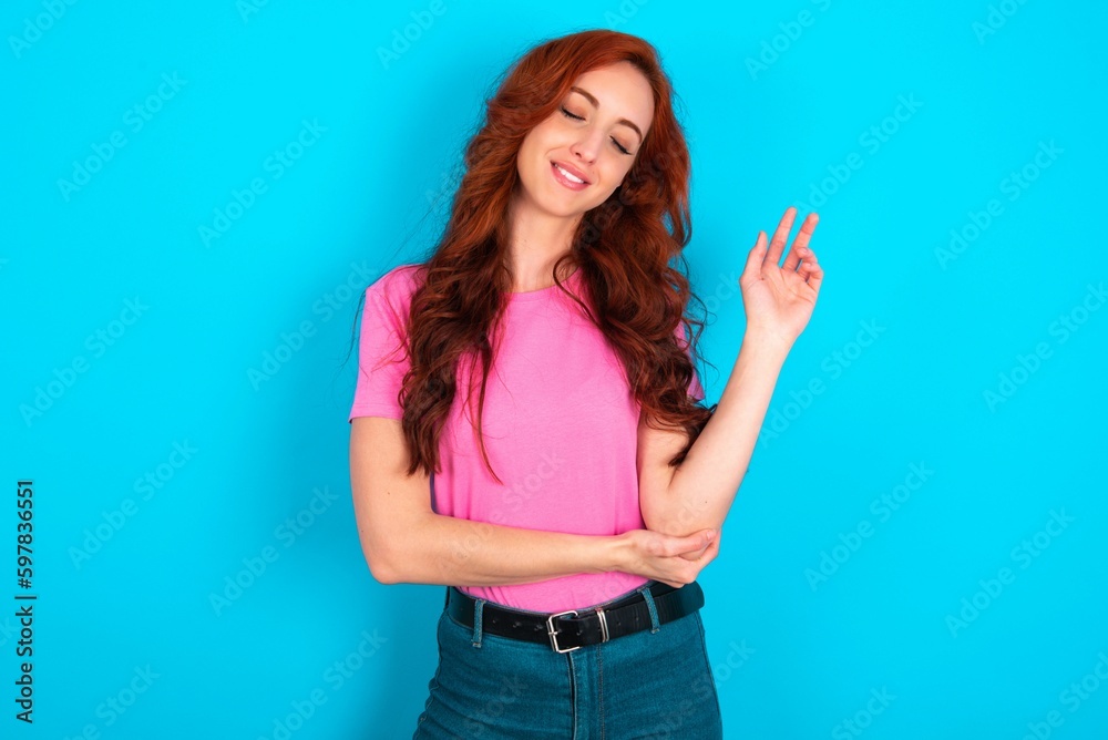 Overjoyed successful young redhead woman wearing pink T-shirt over blue background raises palm and closes eyes in joy being entertained by friends Overjoyed successful young redhead woman wearing pink T-shirt over blue background raises palm and closes eyes in joy being entertained by friends