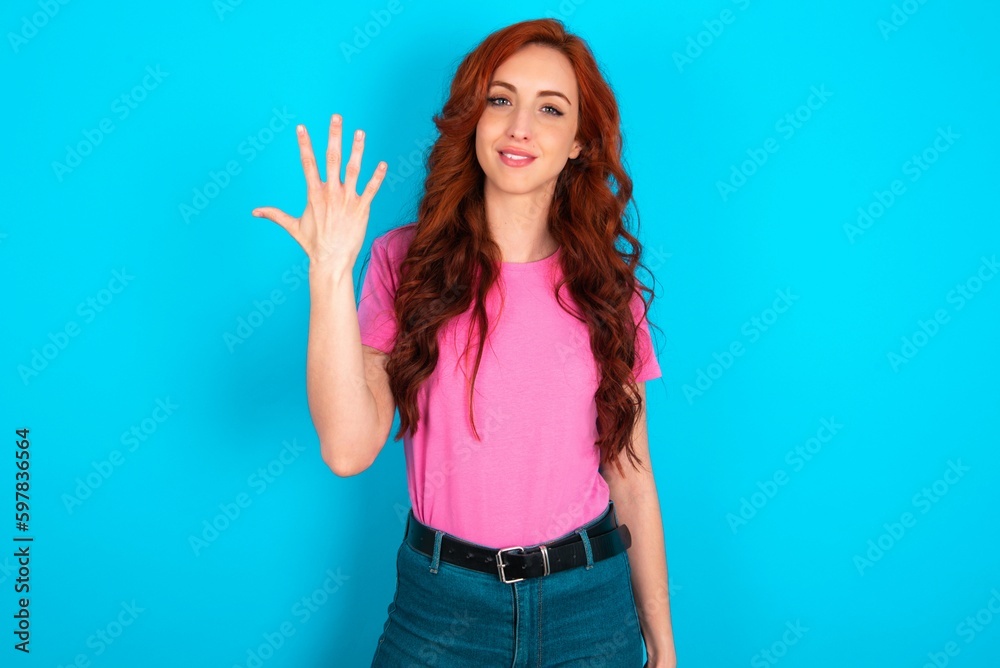 young redhead woman wearing pink T-shirt over blue background smiling and looking friendly, showing number five or fifth with hand forward, counting down young redhead woman wearing pink T-shirt over blue background smiling and looking friendly, showing number five or fifth with hand forward, counting down