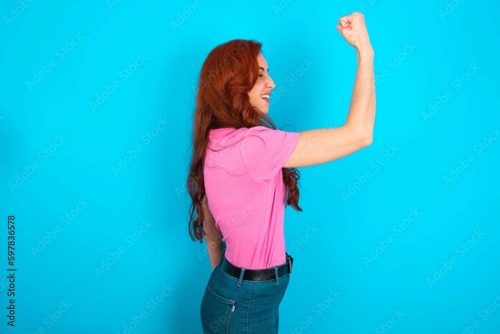 Fototapeta premium Profile photo of young redhead woman wearing pink T-shirt over blue background supporting soccer team World Cup 2022 raise fist shouting