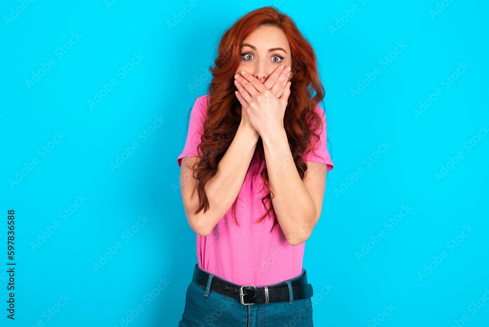Stunned young redhead woman wearing pink T-shirt over blue background covers both hands on mouth, afraids of something astonishing Stunned young redhead woman wearing pink T-shirt over blue background covers both hands on mouth, afraids of something astonishing