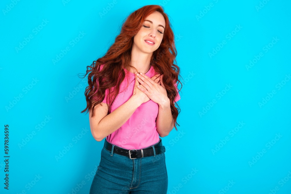 young redhead woman wearing pink T-shirt over blue background expresses happiness, laughs pleasantly, keeps hands on heart young redhead woman wearing pink T-shirt over blue background expresses happiness, laughs pleasantly, keeps hands on heart