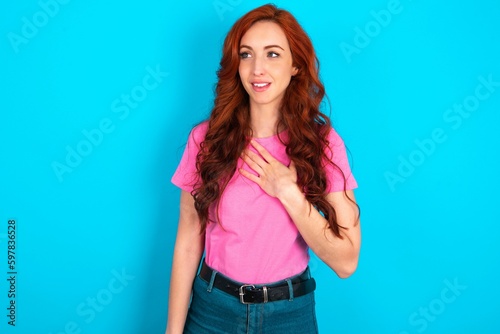Εκτύπωση καμβά Joyful young redhead woman wearing pink T-shirt over blue background expresses positive emotions recalls something funny keeps hand on chest and giggles happily