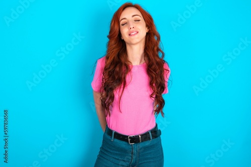 Positive young redhead woman wearing pink T-shirt over blue background with overjoyed expression closes eyes and laughs shows white perfect teeth © Jihan