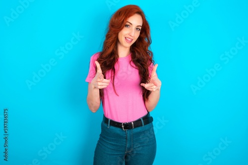 young redhead woman wearing pink T-shirt over blue background directs fingers at camera selects someone. I recommend you. Best choice