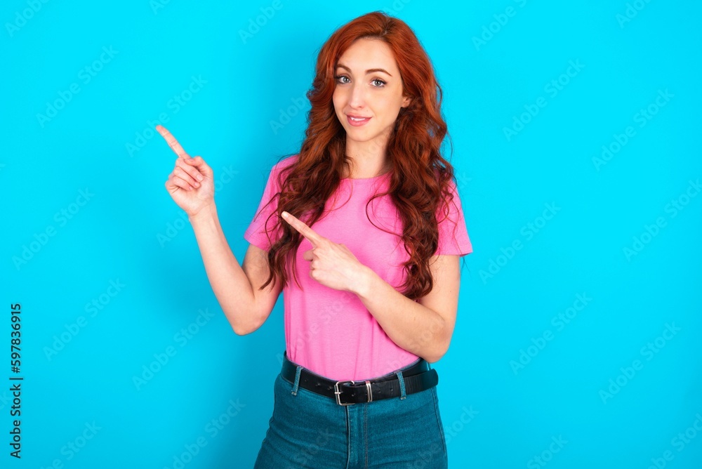 Fototapeta premium young redhead woman wearing pink T-shirt over blue background points at copy space indicates for advertising gives right direction