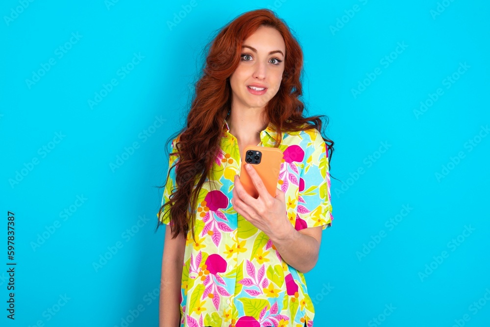 Young redhead woman wearing colorful shirt over blue background holds mobile phone in hands and rejoices positive news, uses modern cellular Young redhead woman wearing colorful shirt over blue background holds mobile phone in hands and rejoices positive news, uses modern cellular