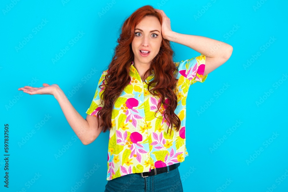 Shocked amazed surprised Young redhead woman wearing colorful shirt over blue background hold hand offering proposition Shocked amazed surprised Young redhead woman wearing colorful shirt over blue background hold hand offering proposition