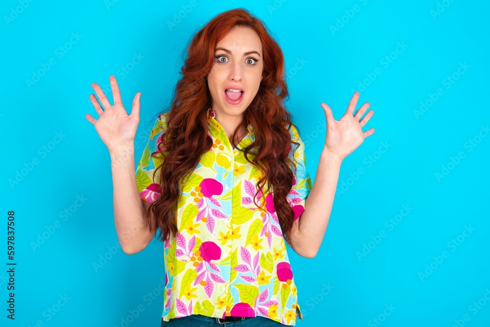 Delighted positive Young redhead woman wearing colorful shirt over blue background opens mouth and arms palms up after having great result Delighted positive Young redhead woman wearing colorful shirt over blue background opens mouth and arms palms up after having great result
