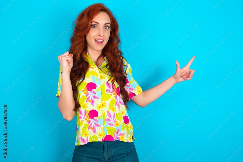 Young redhead woman wearing colorful shirt over blue background points at empty space holding fist up, winner gesture. Young redhead woman wearing colorful shirt over blue background points at empty space holding fist up, winner gesture.