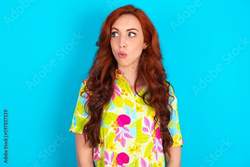 Shocked Young redhead woman wearing colorful shirt over blue background look empty space with open mouth screaming: Oh My God! I can't believe this.