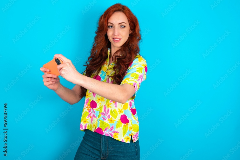 Nice addicted cheerful Young redhead woman wearing colorful shirt over blue background using gadget playing network game Nice addicted cheerful Young redhead woman wearing colorful shirt over blue background using gadget playing network game