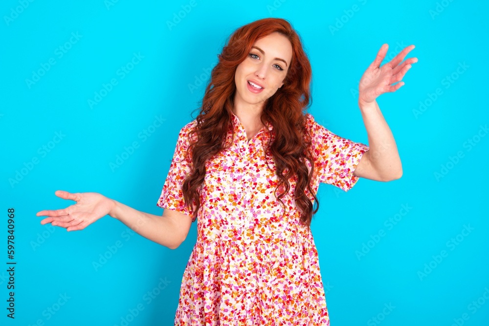 Cheerful Caucasian redhead woman wearing floral dress over blue background making a welcome gesture raising arms over head. Cheerful Caucasian redhead woman wearing floral dress over blue background making a welcome gesture raising arms over head.
