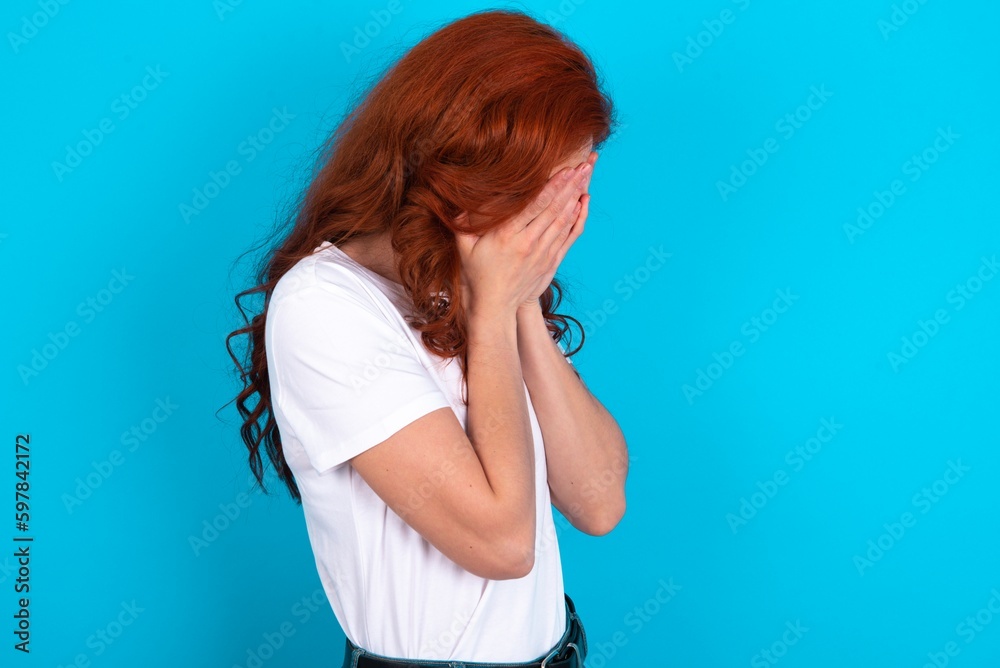 Sad young redhead woman wearing white T-shirt over blue background covering face with hands and crying.