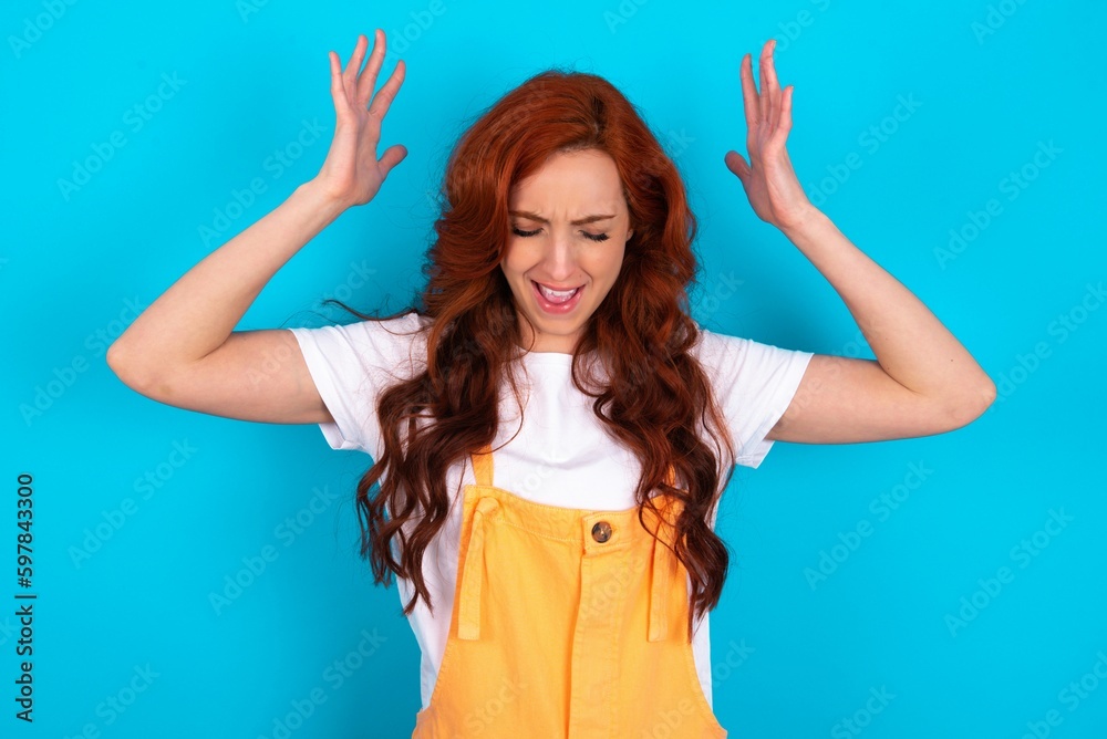 Young redhead woman wearing orange overall over blue background goes crazy as head goes around feels stressed because of horrible situation Young redhead woman wearing orange overall over blue background goes crazy as head goes around feels stressed because of horrible situation