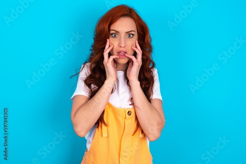Speechless Young redhead woman wearing orange overall over blue background keeps hands near opened mouth reacts to shocking news stares wondered at camera