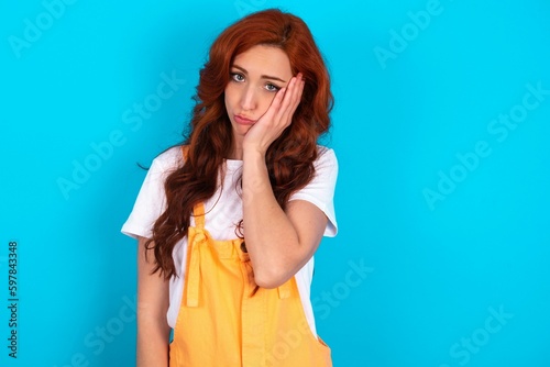 Sad lonely Young redhead woman wearing orange overall over blue background touches cheek with hand bites lower lip and gazes with displeasure. Bad emotions