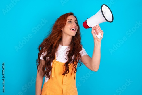 Funny Young redhead woman wearing orange overall over blue background People sincere emotions lifestyle concept. Mock up copy space. Screaming in megaphone.