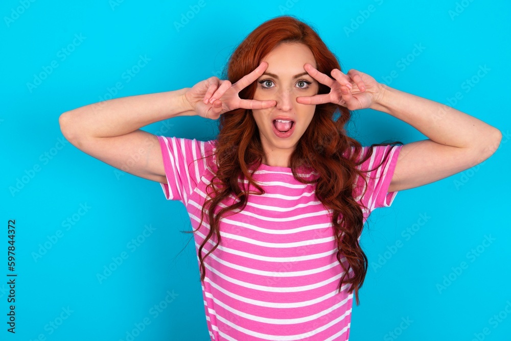 Cheerful positive young redhead woman wearing striped T-shirt over blue background shows v-sign near eyes open mouth