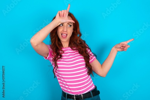 young redhead woman wearing striped T-shirt over blue background showing loser sign and pointing at empty space