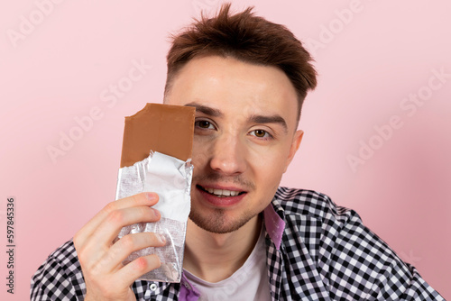A happy young man drinking coke and eating a chocolate bar. fast food and sugar addiction, unhealthy diet. Isolated on pink background.