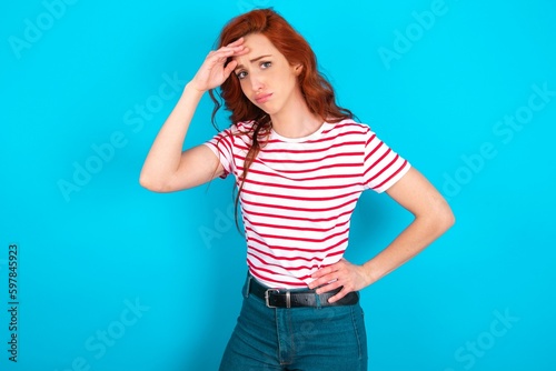 young redhead woman wearing striped T-shirt over blue background having problems, worried and stressed holds hand on forehead.