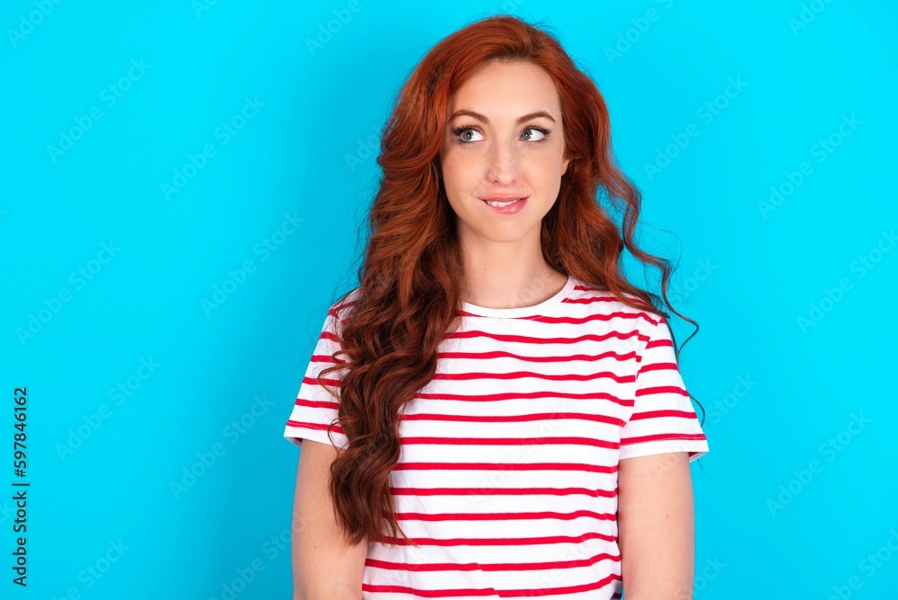 Amazed young redhead woman wearing striped T-shirt over blue background bitting lip and looking tricky to empty space. Amazed young redhead woman wearing striped T-shirt over blue background bitting lip and looking tricky to empty space.