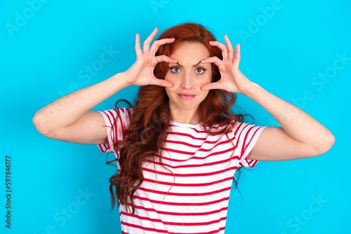 young redhead woman wearing striped T-shirt over blue background keeping eyes opened to find a success opportunity.