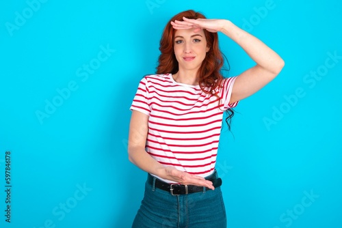 young redhead woman wearing striped T-shirt over blue background gesturing with hands showing big and large size sign, measure symbol. Smiling looking at the camera.