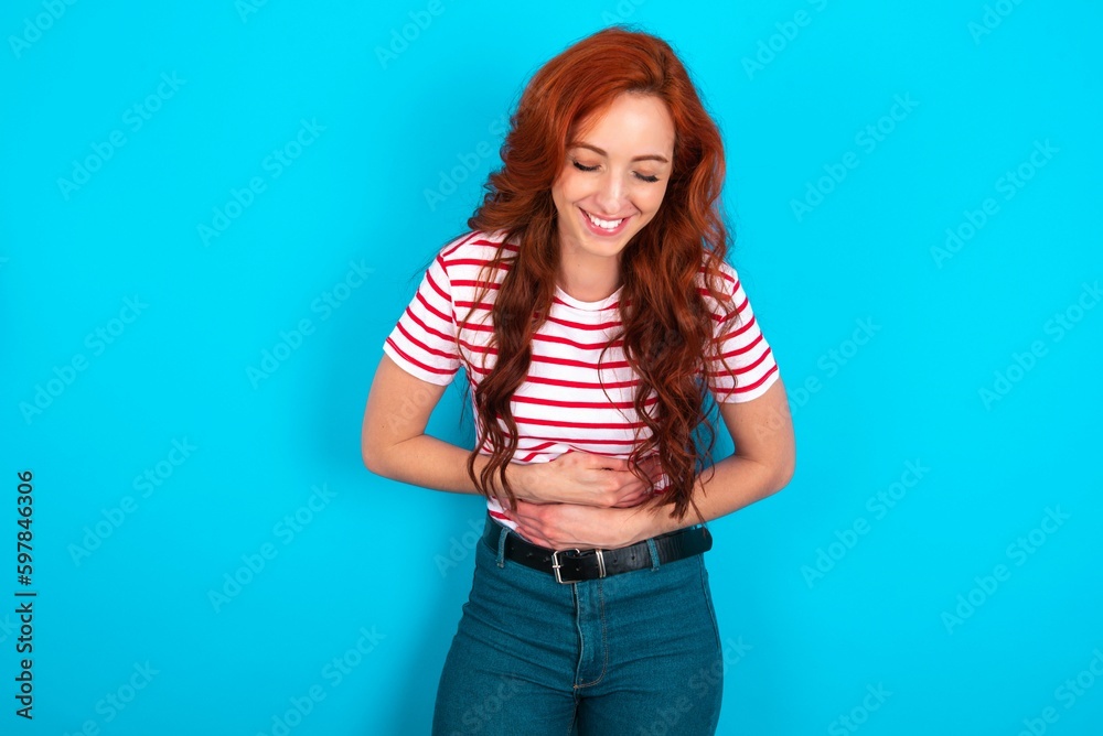 young redhead woman wearing striped T-shirt over blue background smiling and laughing hard out loud because funny crazy joke with hands on body. young redhead woman wearing striped T-shirt over blue background smiling and laughing hard out loud because funny crazy joke with hands on body.
