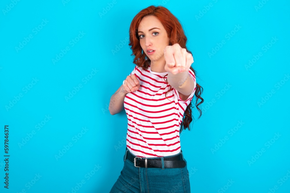 young redhead woman wearing striped T-shirt over blue background Punching fist to fight, aggressive and angry attack, threat and violence young redhead woman wearing striped T-shirt over blue background Punching fist to fight, aggressive and angry attack, threat and violence