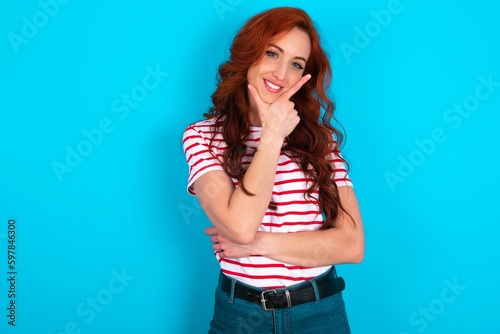 young redhead woman wearing striped T-shirt over blue background looking confident at the camera smiling with crossed arms and hand raised on chin. Thinking positive.
