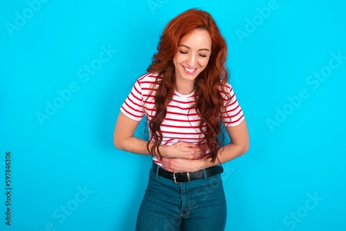 young redhead woman wearing striped T-shirt over blue background smiling and laughing hard out loud because funny crazy joke with hands on body.