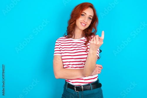 young redhead woman wearing striped T-shirt over blue background Beckoning come here gesture with hand inviting welcoming happy and smiling