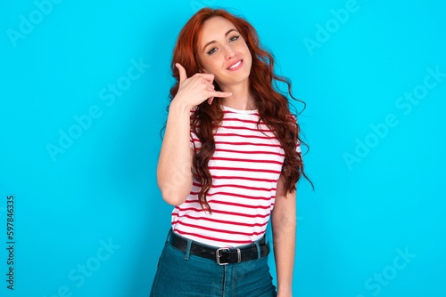 young redhead woman wearing striped T-shirt over blue background smiling doing phone gesture with hand and fingers like talking on the telephone. Communicating concepts.