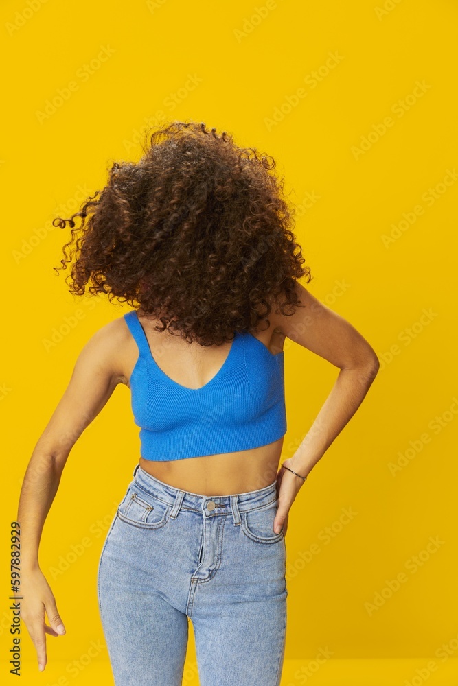 Woman with curly afro hair in a blue T-shirt on a yellow background dancing flying hair with sunglasses yellow, hand signs, look into the camera, smile with teeth and happiness, copy space Woman with curly afro hair in a blue T-shirt on a yellow background dancing flying hair with sunglasses yellow, hand signs, look into the camera, smile with teeth and happiness, copy space