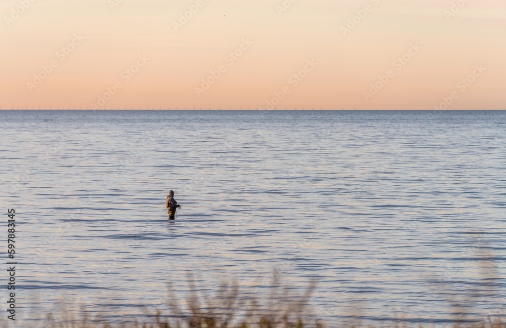 Fototapeta premium An angler in waterproof trousers is fishing in the Baltic Sea. He is standing on a stone in the water. 