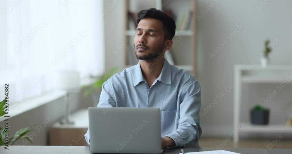 Pensive Indian freelancer guy working on laptop seated at workplace desk in homeoffice, feels puzzled, search creative ideas and business solutions, doing on-line telework at home using Generative AI