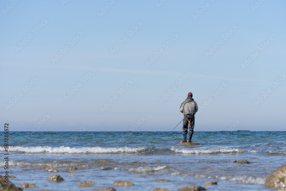An angler in waterproof trousers is fishing in the Baltic Sea. He is standing on a stone in the water. An angler in waterproof trousers is fishing in the Baltic Sea. He is standing on a stone in the water.