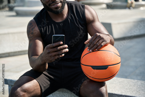 Fit sporty young African black ethnic man sitting outdoors holding basketball ball and mobile phone using apps, looking at smartphone, resting after street sport game with cellphone. Closeup