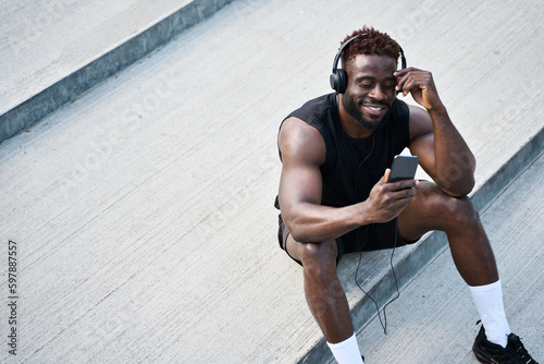 Strong fit sporty young black man sitting on urban stairs holding phone using mobile apps listening music. Strong African guy wearing headphones looking at smartphone outdoors. Top view, copy space